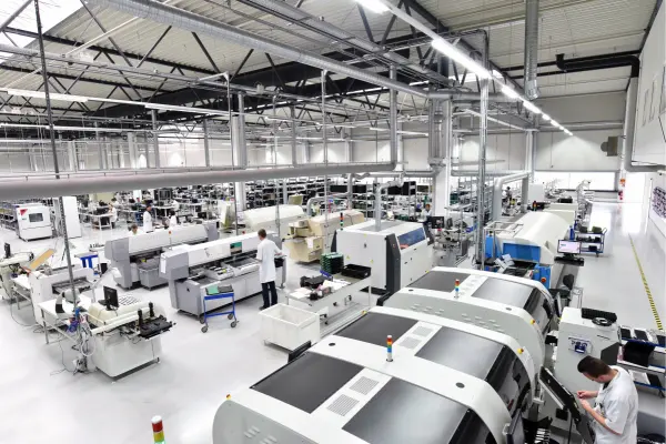 A large, modern manufacturing facility with workers in white coats operating advanced machinery and equipment under a high ceiling.